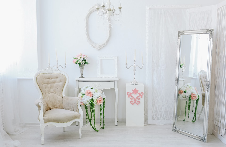 Angolo di un salotto bianco in stile shabby, con poltrona antica, lampadario a candelabro, grande specchio a terra, decorazioni floreali, consolle e cornici vuote decorative