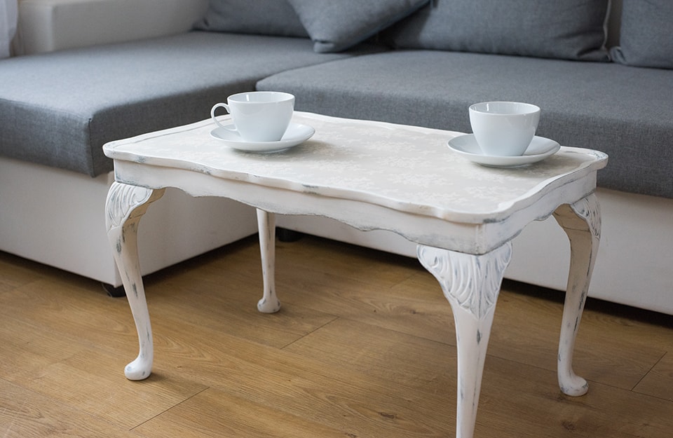 Tavolino da caffè bianco in stile shabby chic, con sopra due tazze. Sullo sfondo si vede parte di un sofà moderno