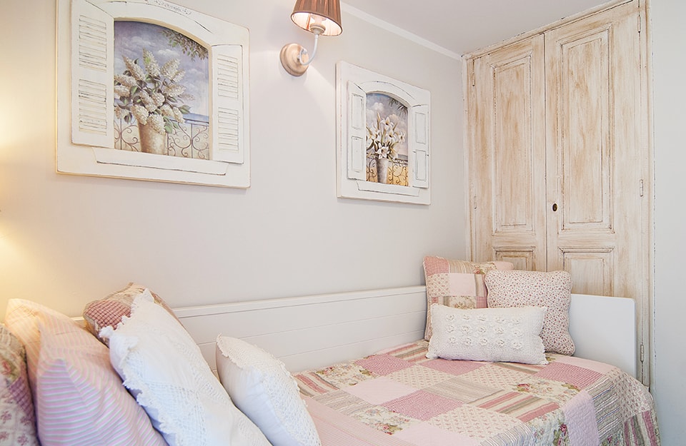 Camera da letto romantica con coperta patchwork rosa, quadri trompe-l'oeil che simulano delle finestre con vasi fioriti e un vecchio armadio restaurato in stille shabby chic