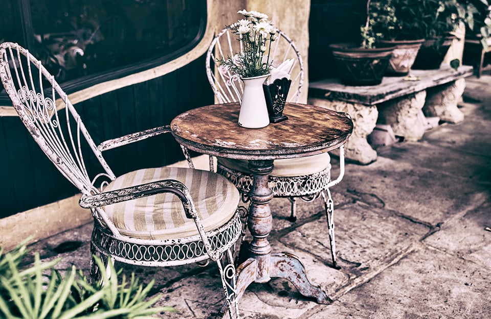 Ambiente esterno con tavolino e due sedie in stile shabby chic. Sul tavolo ci sono un vaso con fiori e un porta-tovaglioli