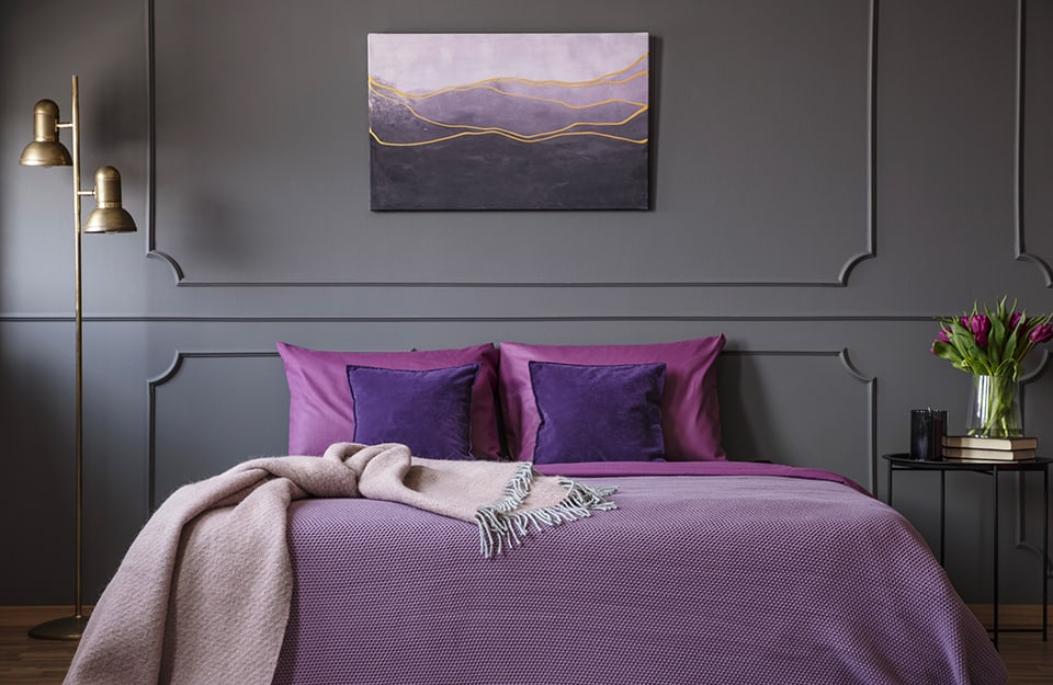 Camera da letto con pareti scure e decorate. La biancheria è sulle tonalità del viola, così come un quadro appeso sopra la testiera