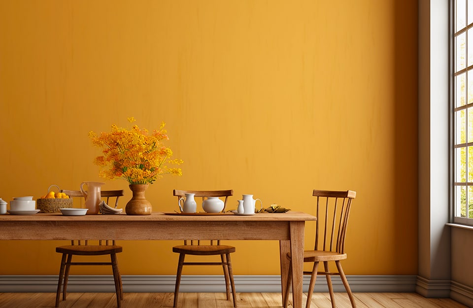 Sala da pranzo con parete color senape, tavolo (apparecchiato) e sedie in stile nordico in legno naturale, vaso con fiori gialli