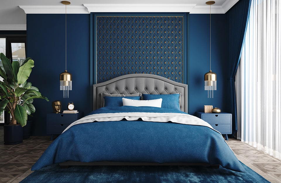 Una stanza da letto con pareti color blu cobalto e un letto matrimoniale con biancheria dello stesso colore, testiera grigia e, dietro, una decorazione in stile art déco. Accanto al letto ci sono due comodini blu, sui quali scendono delle lampade tondeggianti metalliche color oro, con pendant in cristallo