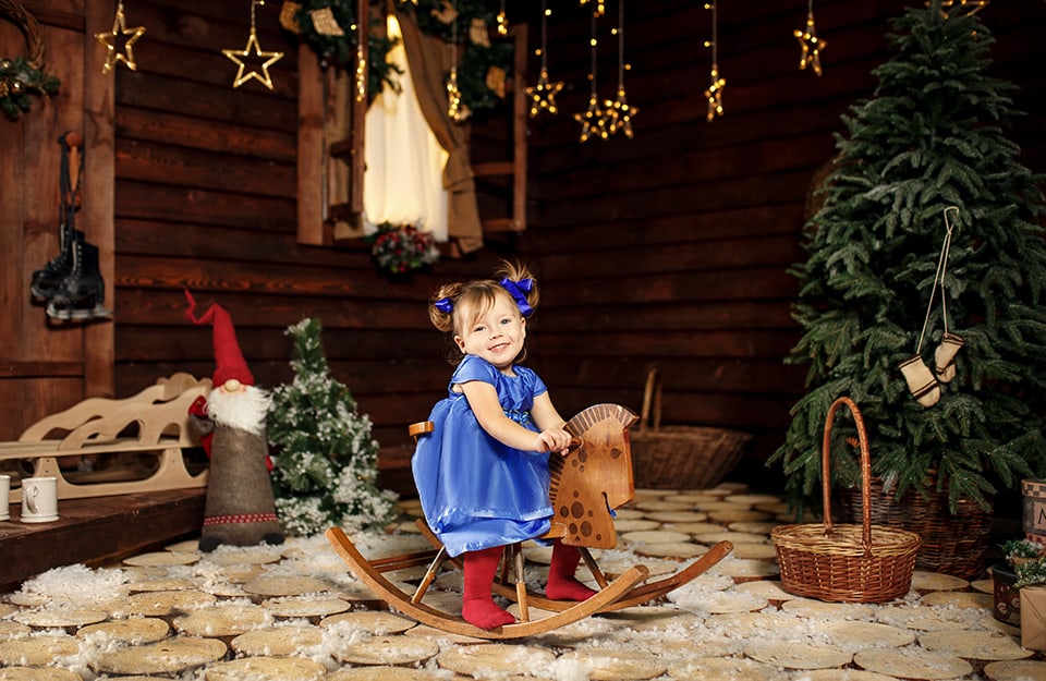 Bambina piccola con vestitino blu e fiocchetti ai capelli su cavallino a dondolo in legno in una stanza molto natalizia in stile baita di montagna