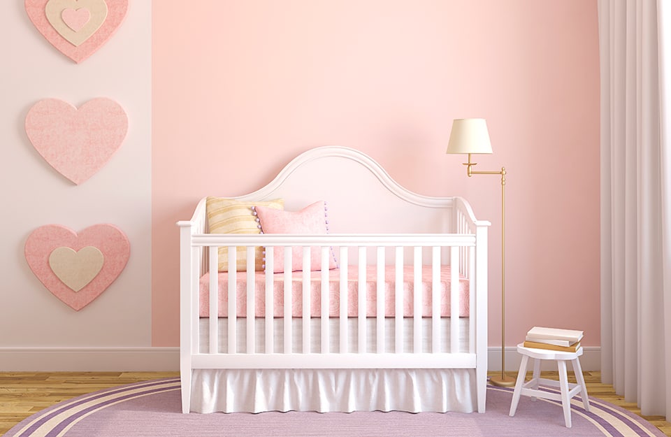 Cameretta per una neonata con culla, seggiolina e lampada. Pareti e tappeto sono rosa, così come le decorazioni a cuore appese e le coperte della culla