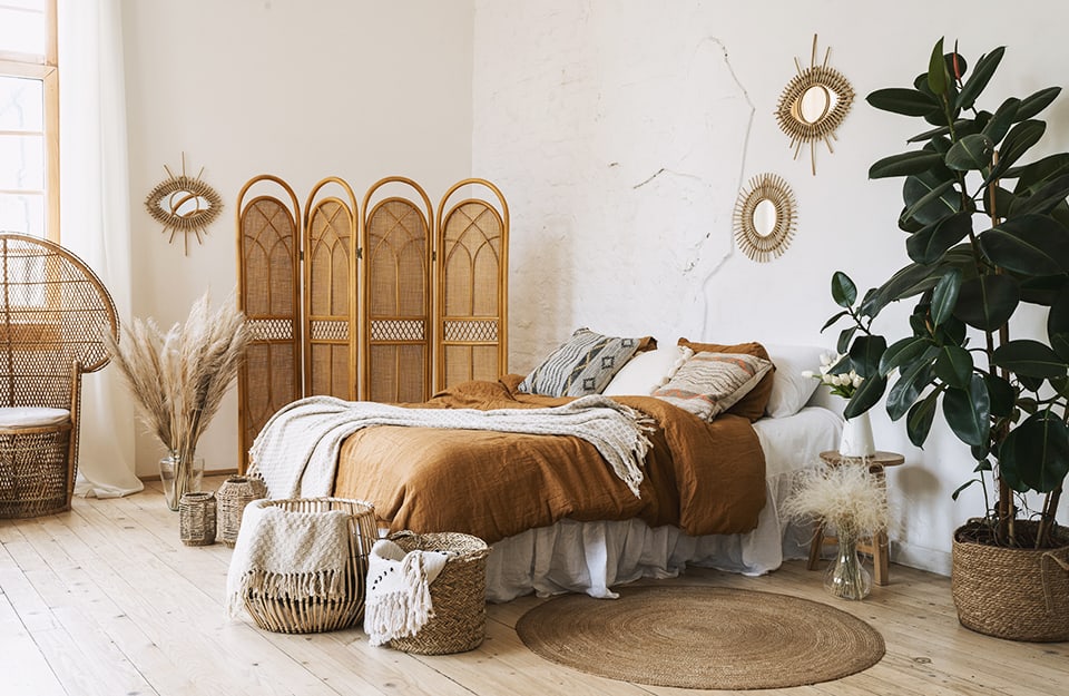 Camera da letto in stile Boho Chic con tanti elementi in vimini e rattan, specchi a forma di occhio, piante e coperte dai colori caldi
