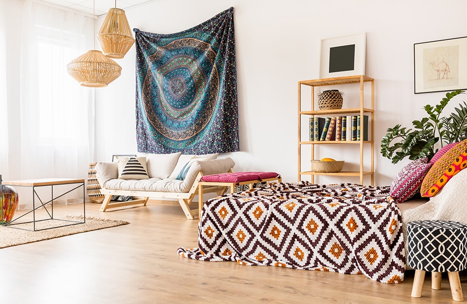 Open space Boho Chic, con divano in stile scandinavo, libreria minimale, lampadari in bambù intrecciato, coperte e cuscini a pattern etnici, una grande telo d'ispirazione hippy appeso alla parete.