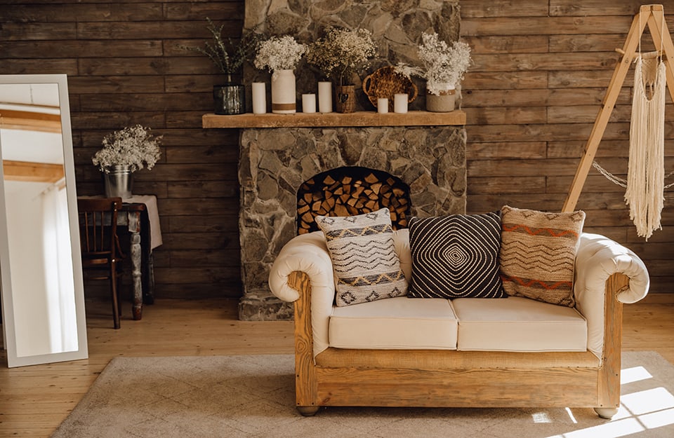 Salotto confortevole e caldo, stile cottagecore. Un divano di legno con cuscini rustici, un camino pieno di legna, molti soprammobili