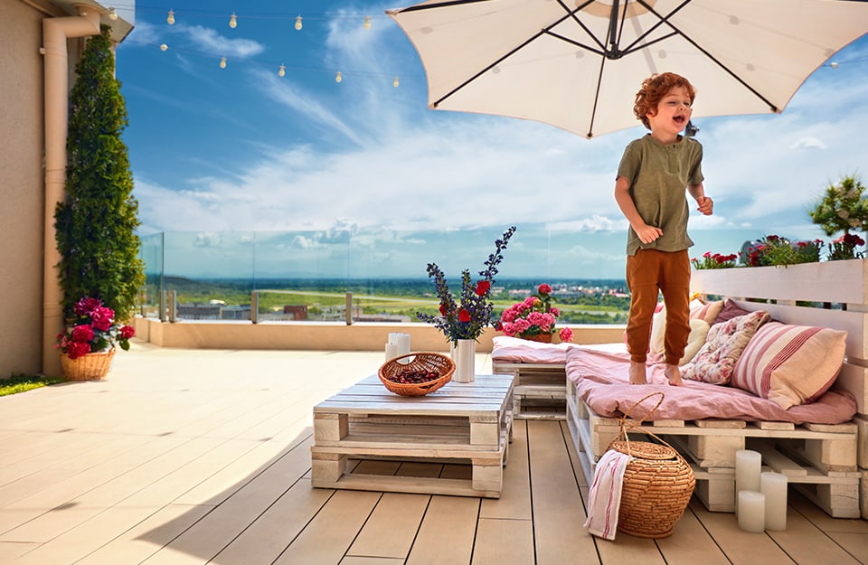 Grande terrazza dove un bambino, all'ombra di un ombrellone, gioca in piedi su un divano costruito coi pallet, con cui è stato realizzato anche un tavolino da caffè.