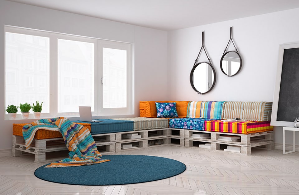 Salotto luminoso e spazioso con grande divano d'angolo realizzato coi pallet. I cuscini sono multicolore. Nella stanza si vedono anche una lavagna appoggiata al muro e due specchi circolari appesi