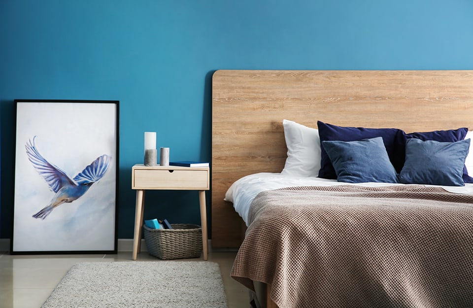 Camera da letto con parete azzurra, letto e comodino in legno e un quadro appoggiato a terra con l'immagine di un uccello in volo