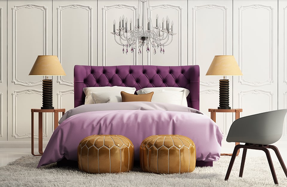Camera da letto in stile vintage con pareti decorate bianche, lampadario a candelabro sopra il letto, testiera viola con impunture e biancheria sui toni del lilla