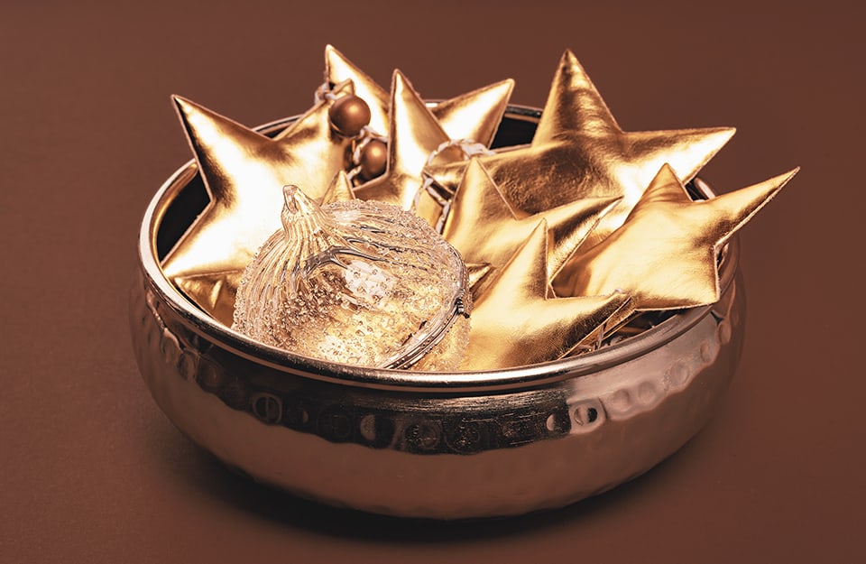 Centrotavola natalizio con ciotola metallica riempita di stelle dorate in tessuto e una pallina di vetro, il tutto su sfondo marrone monocromatico