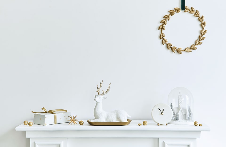 Composizione natalizia di soprammobili e decorazioni in total white e vernici metallizzate: ci sono un cervo, un pacco regalo, una sveglia, una sfera di neve e, appena, una ghirlanda di foglie