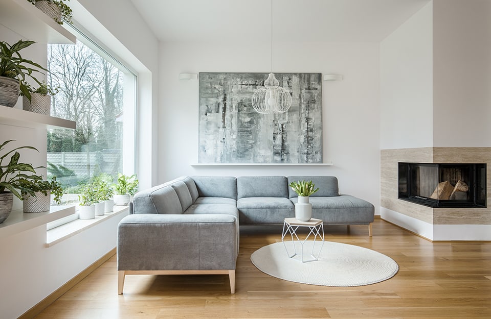 Salotto minimale con divano in stile scandinavo, camino squadrato d'angolo, un grande quadro che domina la parete bianca, pavimento a parquet, mensole piene di piante e una grande finestra orizzontale che dà su un giardino