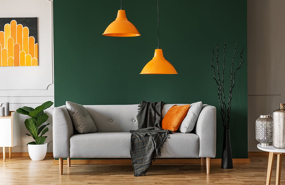 Salotto con colori a contrasto tra verde (parete e piante) e arancio (quadro, lampadari e cuscino), esaltati dall'uso di altri colori, come il sofà grigio, la coperta e i cuscini neri