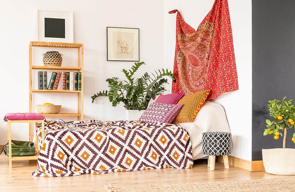 Una camera da letto minimale arricchita da tocchi in stile etnico grazie a un telo appeso alla parete, dei cuscini con stampe geometriche, una stampa con un cammello, delle piante e dei cesti in vimini