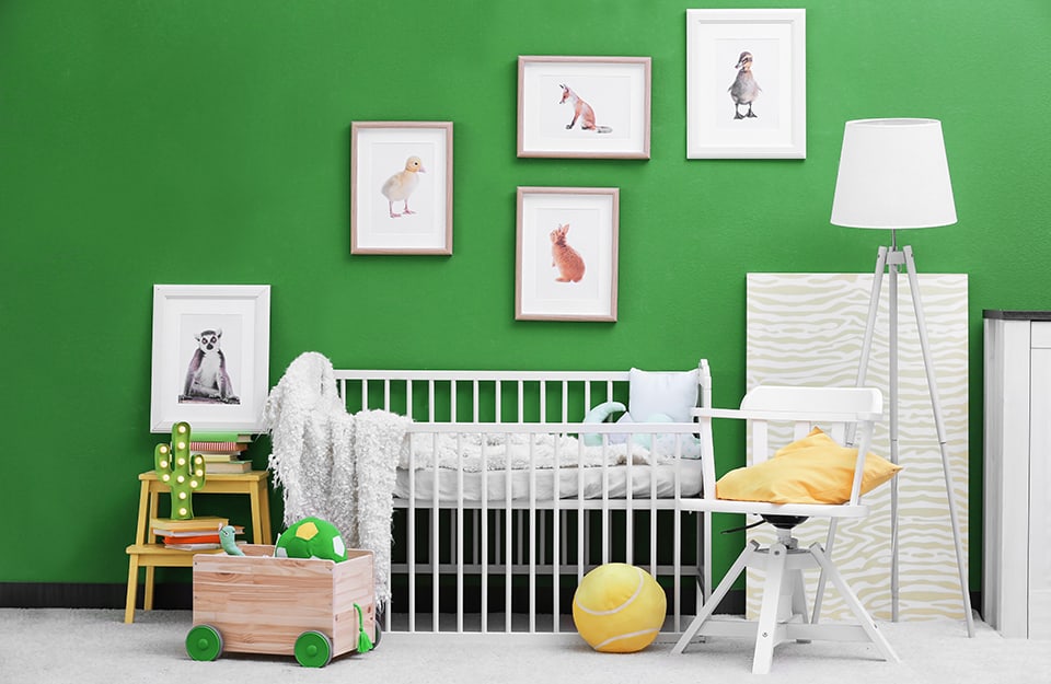Cameretta per bambino con lettino, giochi, sedia, lampada a piedistallo, libri, sgabello e disegni incorniciati appesi a una parete color “zoomer green”