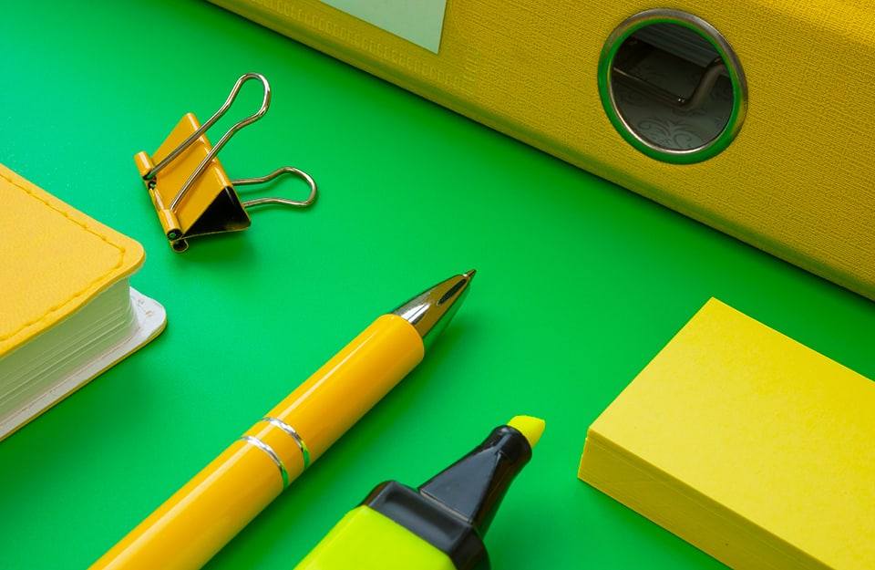 Primo piano di strumenti da cancelleria (agenda, penna, evidenziatore, post-it, raccoglitore e clip) su una scrivania col verde acceso, o “zoomer green”