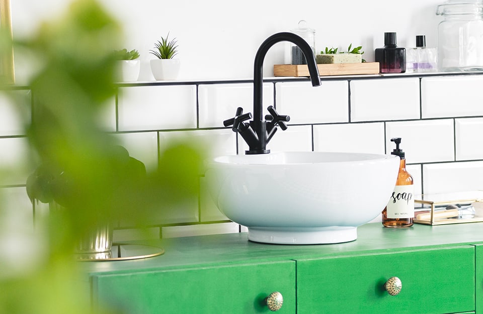 Dettaglio di una stanza da bagno con piastrelle bianche, lavabo bianco, rubinetteria nera e mobiletto verde acceso
