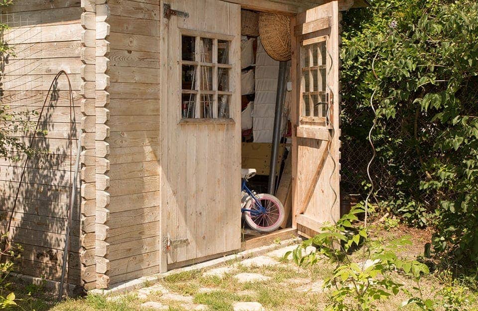 Dettagli di una casetta in legno da giardino con la porta aperta, dalla quale si vedono attrezzi e una piccola bici da bambini