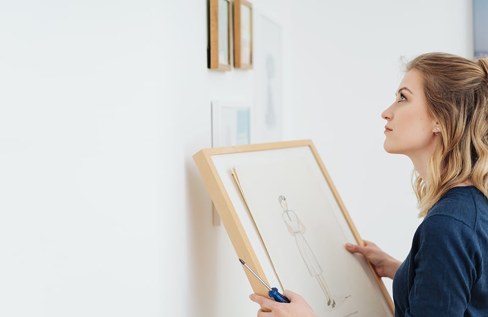 Una donna bionda sta creando una composizione di quadri alla parete, ha un quadro in mano e sta pensando a dove attaccarlo