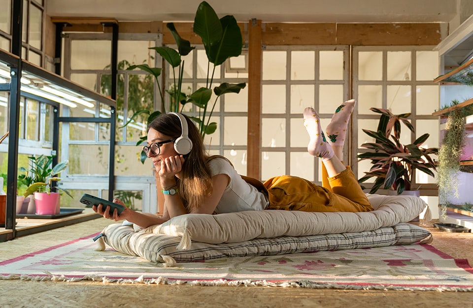 Una ragazza con cuffie e smartphone ascolta un podcast sdraiata su dei cuscini in uno spazio decorato in stile industriale
