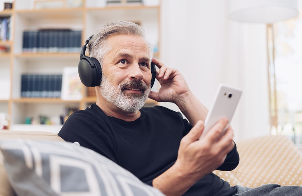 Un uomo giovanile con barba e capelli bianchi sta ascoltando un podcast con cuffie e smartphone seduto sul divano