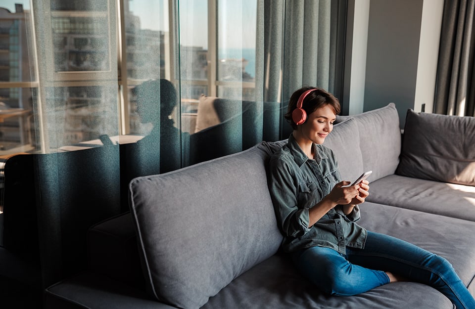 Una ragazza ascolta un podcast con cuffie e smartphone seduta su un divano davanti a una luminosa finestra che dà, come vediamo dal riflesso, su una grande città