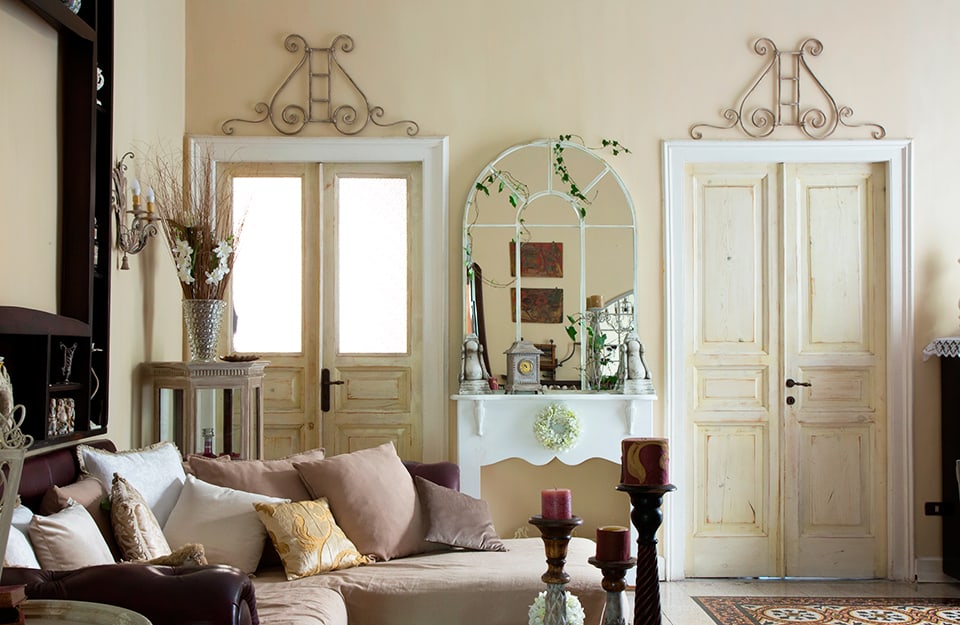 Parte di un salotto in stile provenzale, con due porte, decorazioni in ferro battuto, molti vasi, uno specchio, mobili vintage e un sofà con molti cuscini