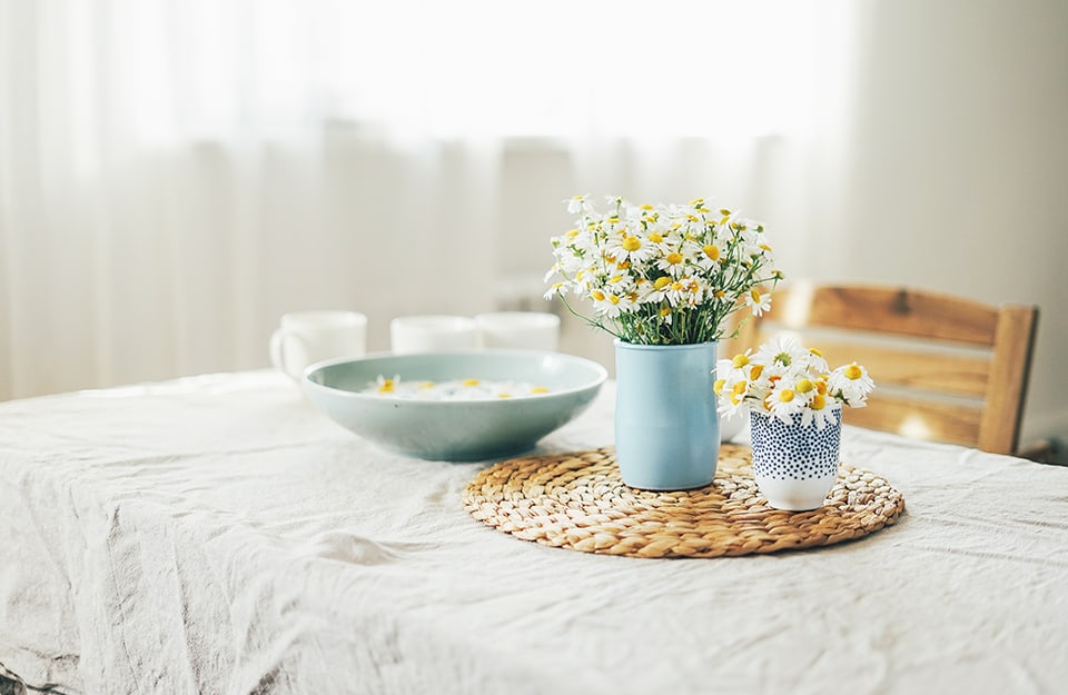 Una luminosa sala da pranzo con una vista d'angolo su una tavola con tovaglia, vasetti di fiori di campo recisi e una ciotola contenente margherite che galleggiano sull'acqua