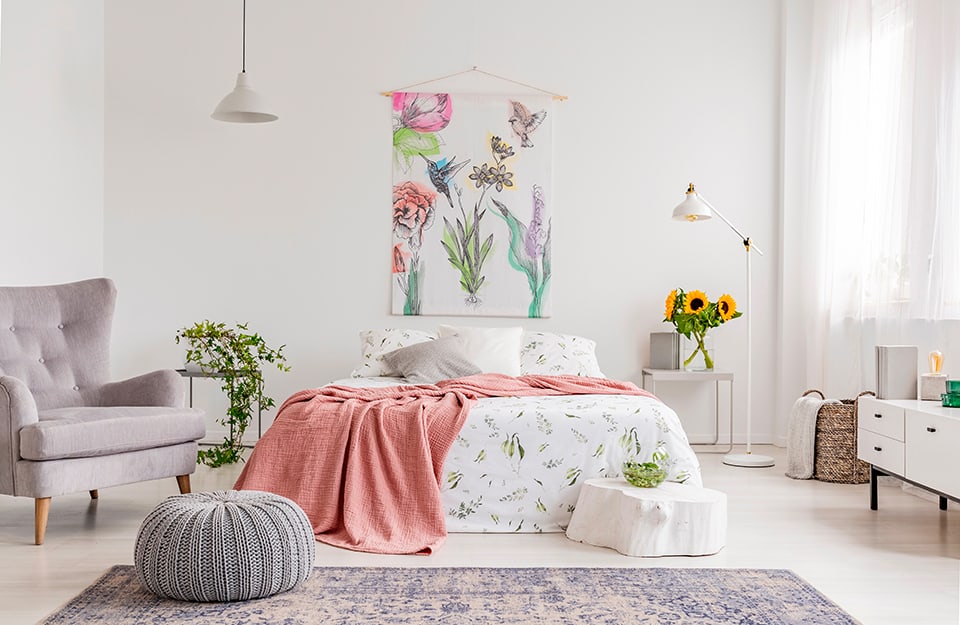 Camera da letto luminosa e sulle tonalità del bianco e del grigio, con molti elementi floreali: un vaso di girasoli, un quadro con dei fiori, lenzuola bianche con stampe a fiori, copriletto rosa pastello