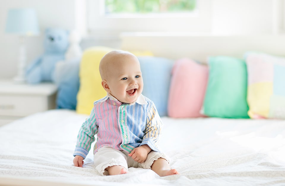 Bambino molto piccolo che ride seduto sopra un letto decorato con diversi cuscini pastello i cui colori richiamano la tutina del piccolo