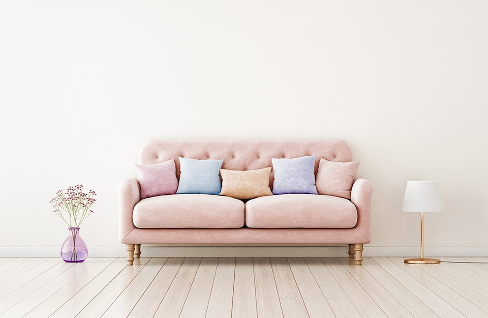 Stanza vuota con sofà trapuntato rosa pastello con sopra cuscini di vari colori pastello e, ai due lati, una lampada da tavolo con paralume bianco e un vaso di vetro lilla con dei fiori