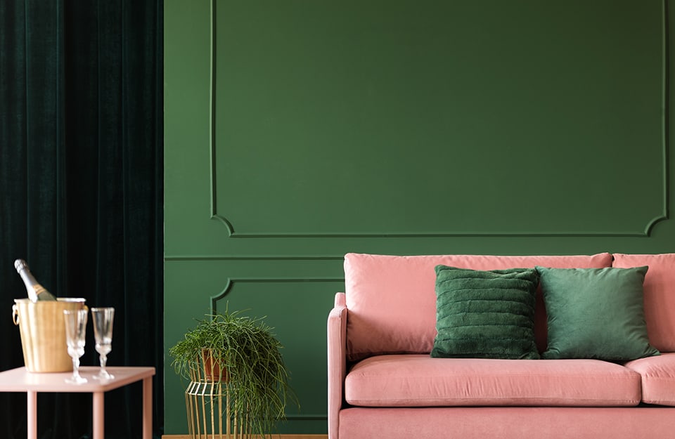 Salotto con pareti verdi decorate con cornici, tavolino da caffè con champagne e bicchieri, una pianta decorativa e un sofà rosa pastello con cuscini verdi