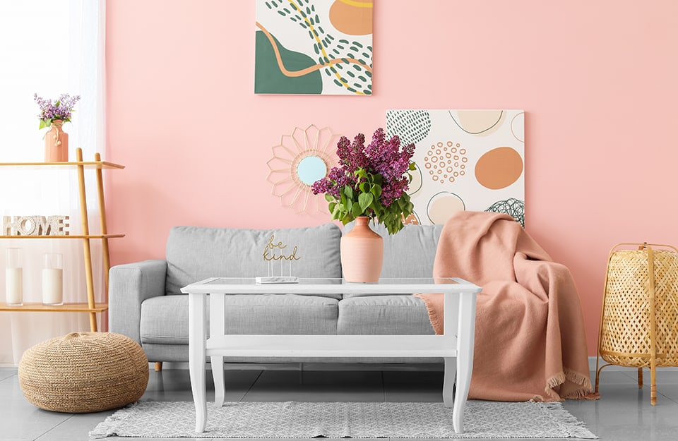 Salotto moderno con pareti rosa pastello, che richiamano il colore del vaso sul tavolino da caffè bianco. Il sofà è grigio chiaro. Ci sono due stampe colorate al muro e due elementi decorativi in vimini