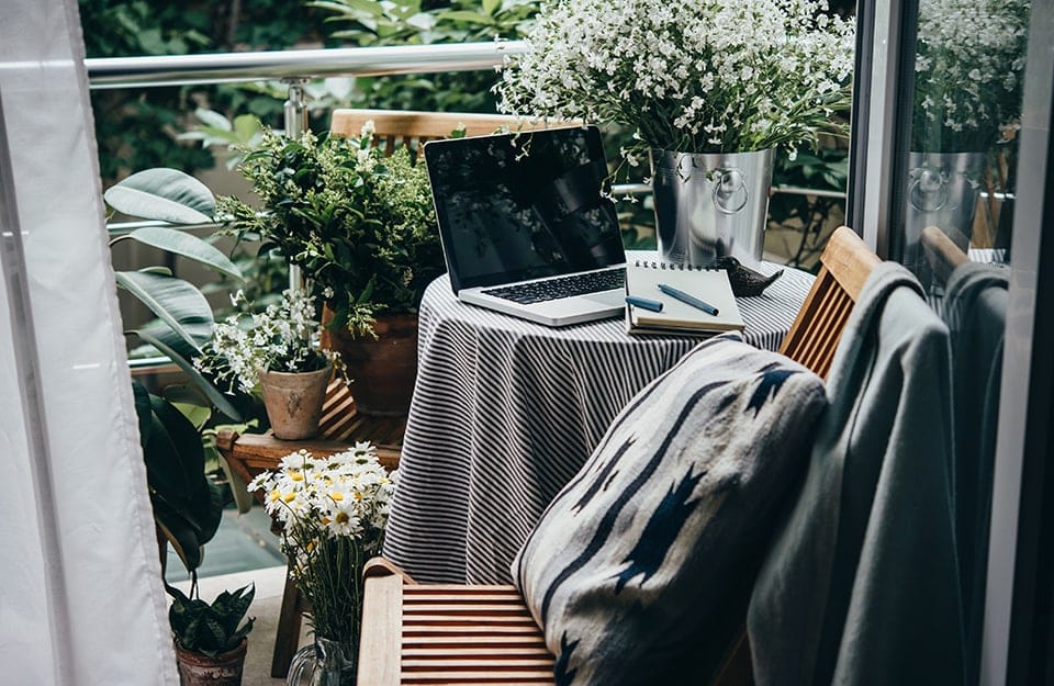 Un tavolino con un computer portatile aperto e un blocco note, su un balcone pieno di piante in vaso e con un paio di sedie in legno