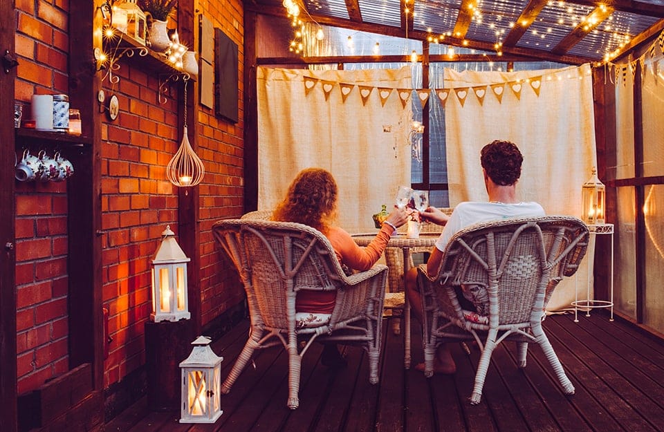 Una coppia brinda seduta a un tavolo, la sera, in una veranda in legno illuminata da file di lampadine e da lanterne