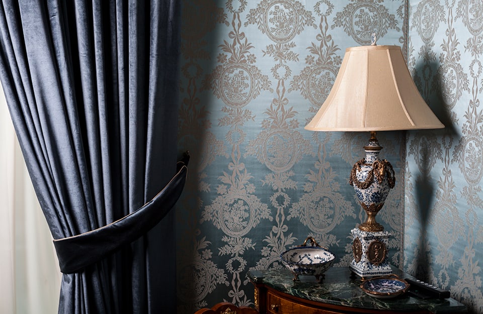 Angolo di una stanza barocca, con carta da parati effetto broccato, abat-jour molto decorata con coprilampada e tendaggio pesante sul blu