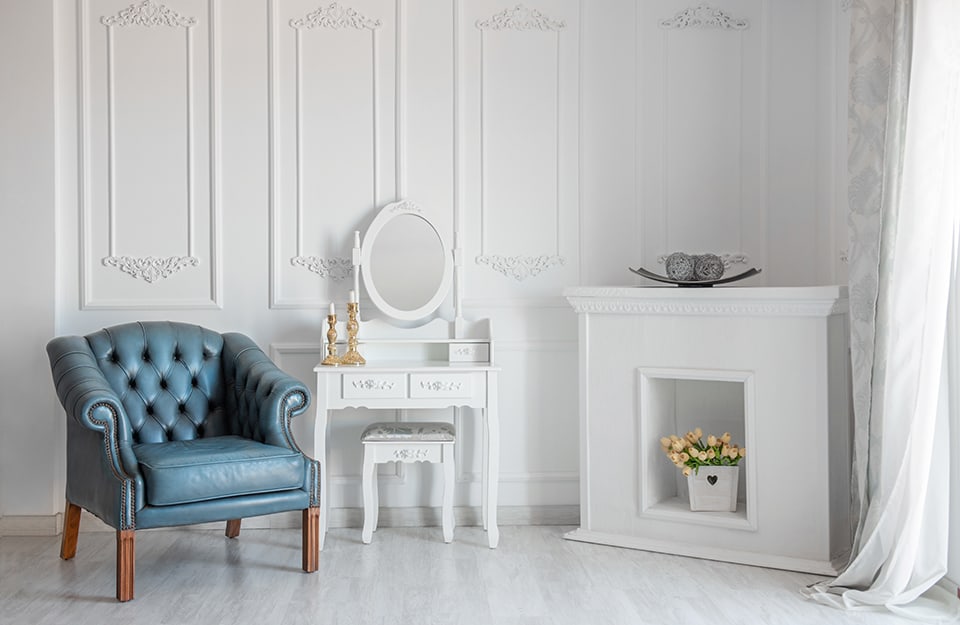 Angolo di salotto in stile barocco moderno, con pareti bianche decorate, pavimento effetto marmo, poltrona blu trapuntata, camino bianco finto, toletta bianca con specchio