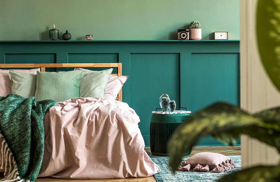 Scorcio su una camera da letto con parete e boiserie sul verde. Il letto è minimale e in legno naturale, con biancheria di vari colori, dal verde al rosa
