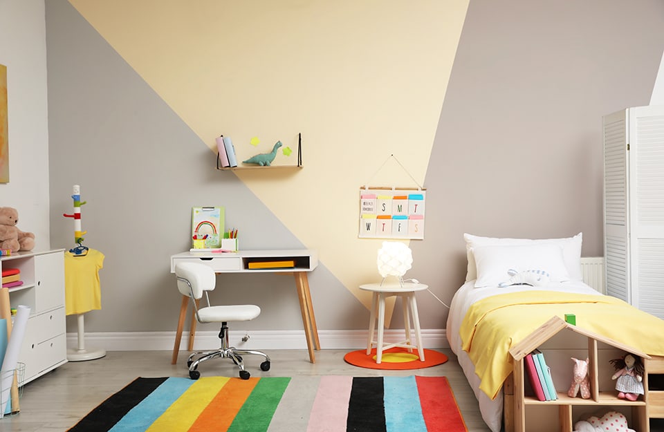 Cameretta dei bambini con parete d'accento beige e triangolo giallo. I mobili sono essenziali e in stile nordico e c'è un tappeto multicolore