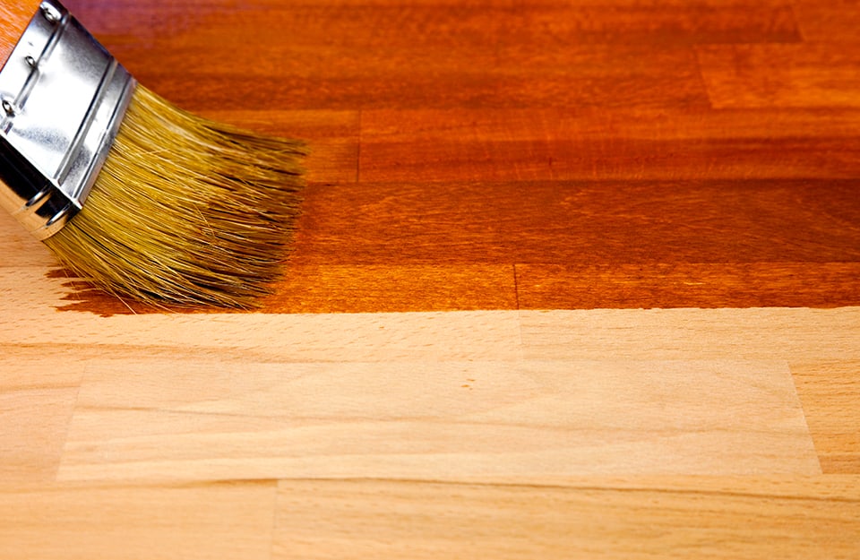 A brush is passing wood stain over a wooden surface and one can clearly see the difference between the darker colour of the already treated surface and the raw, as yet untreated wood;