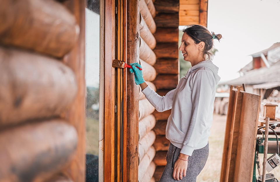 Una mujer trata con impregnante el marco de madera de una ventana en el exterior de una casa toda de madera;