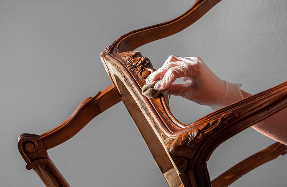 Una mano femminile sta trattando con un tampone imbevuto di mordente il telaio di un'antica sedia in legno che sta restaurando