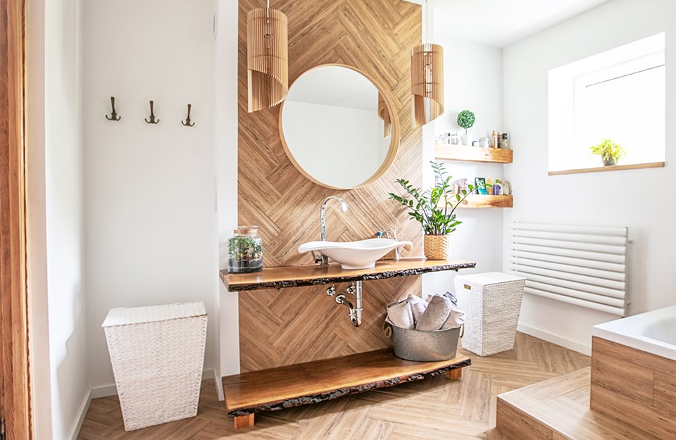 Luminosa stanza da bagno arredata in stile boho chic sui toni del bianco e del legno naturale, con specchio circolare