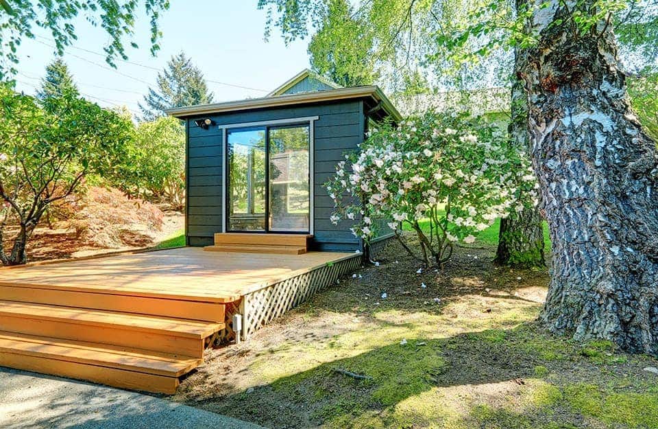 Un giardino rigoglioso con una casetta in legno, sopra una pedana pure in legno, nella quale è stato allestito un garden office