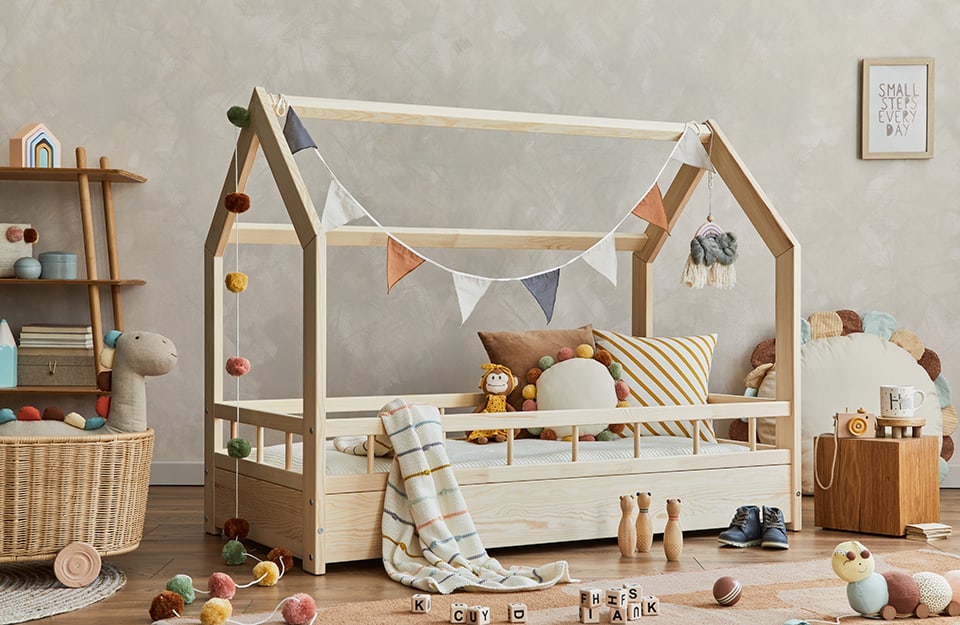 Children's bedroom with bed with wooden frame in the shape of a small house. Coloured pompoms and triangular flags of different colours hang from the frame. The wall is grey. On the floor is a parquet floor with several scattered toys. Next to the bed is a small cubic wooden table with toys on it, a flower-shaped cushion leaning vertically against the wall, a wicker basket with wheels and toys inside, and a small wooden bookcase;