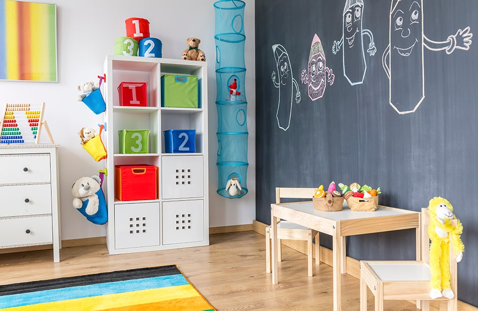 Children's room with a chest of drawers, an Ikea bookcase, toys on the shelves, a small wooden table, parquet flooring on the floor and a wall painted with chalkboard paint with anthropomorphic pencil strokes drawn on it;