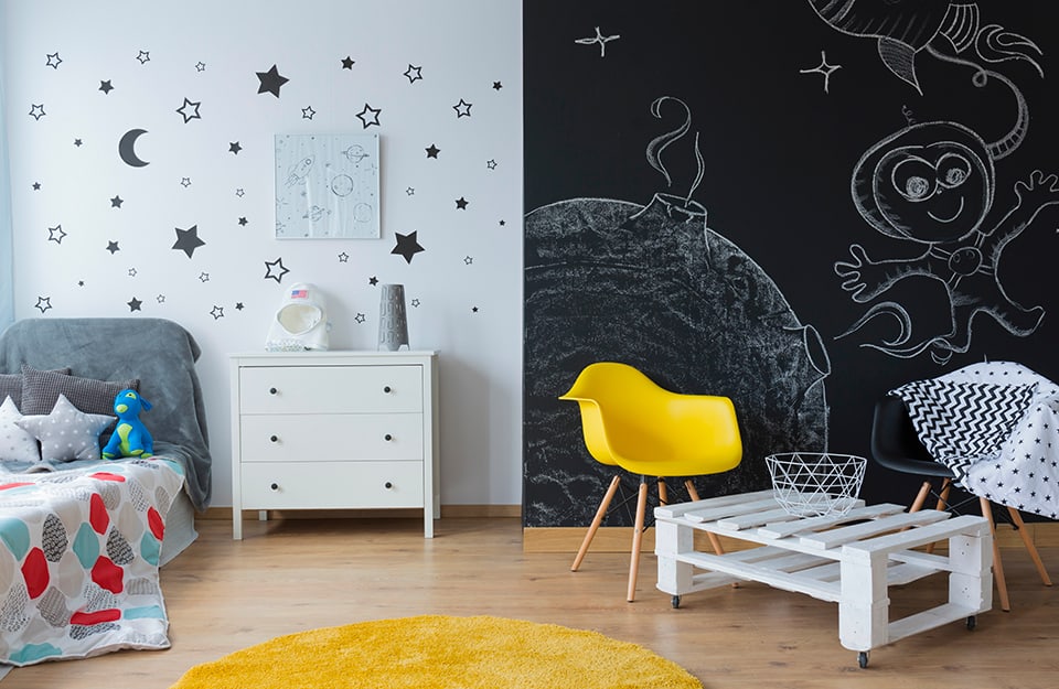 A children's room with a wall divided in half: the left side, painted white, is decorated with stars and moons and there are a bed and a white chest of drawers; the right side of the wall is painted with chalkboard paint and there are drawings on the wall, in front of which stands a modern yellow chair and a coffee table made from two white-painted pallets;
